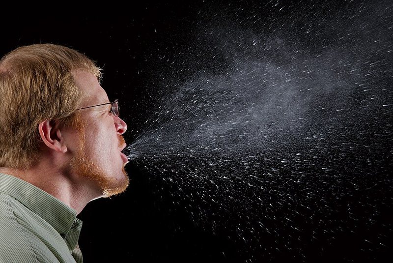 A person sneezing in profile against a black background, with a spray of droplets fanning out from the mouth